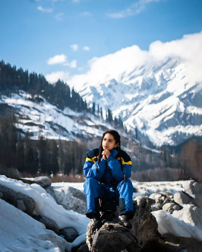 woman in blue jacket and blue backpack sitting on rock near snow covered mountain during daytime